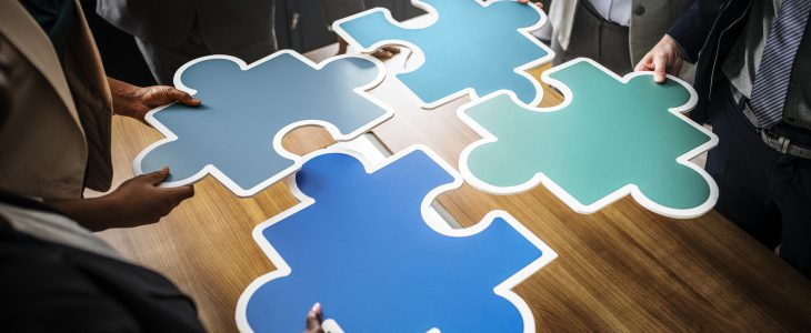 Four people hold together four large green and blue jigsaw puzzle pieces.